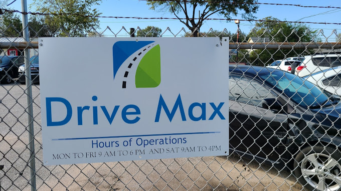 A Drive Max sign on a chain-link fence lists hours of operation: Mon-Fri 9 AM to 6 PM, Sat 9 AM to 4 PM. Parked cars are visible in the background—a snapshot perfect for a Drive Max case study.