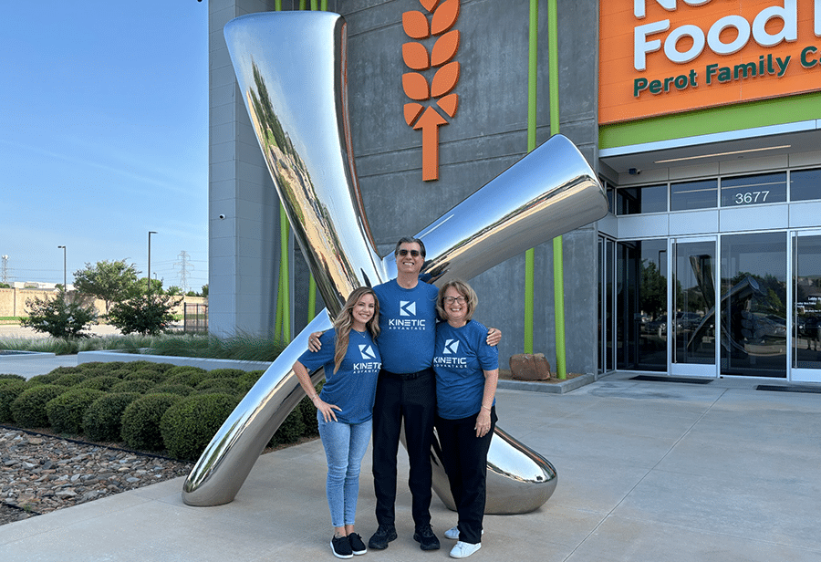 Three people wearing matching blue shirts stand in front of a large metallic sculpture outside the modern North Texas Food Bank building, embodying the spirit of Drive to Thrive.