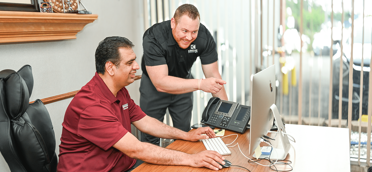 Two men in work uniforms sit and stand at an office desk, looking at a desktop computer screen, with one man pointing and smiling.