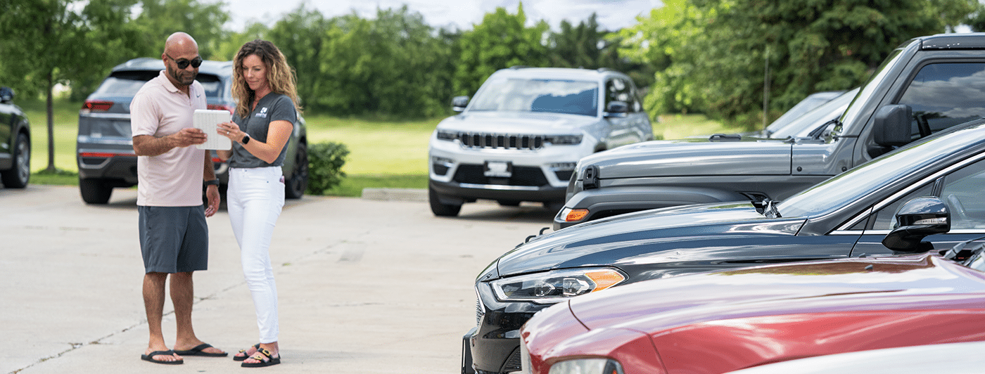 Two people stand in a parking lot looking at a tablet, with several parked cars visible in the foreground and background.